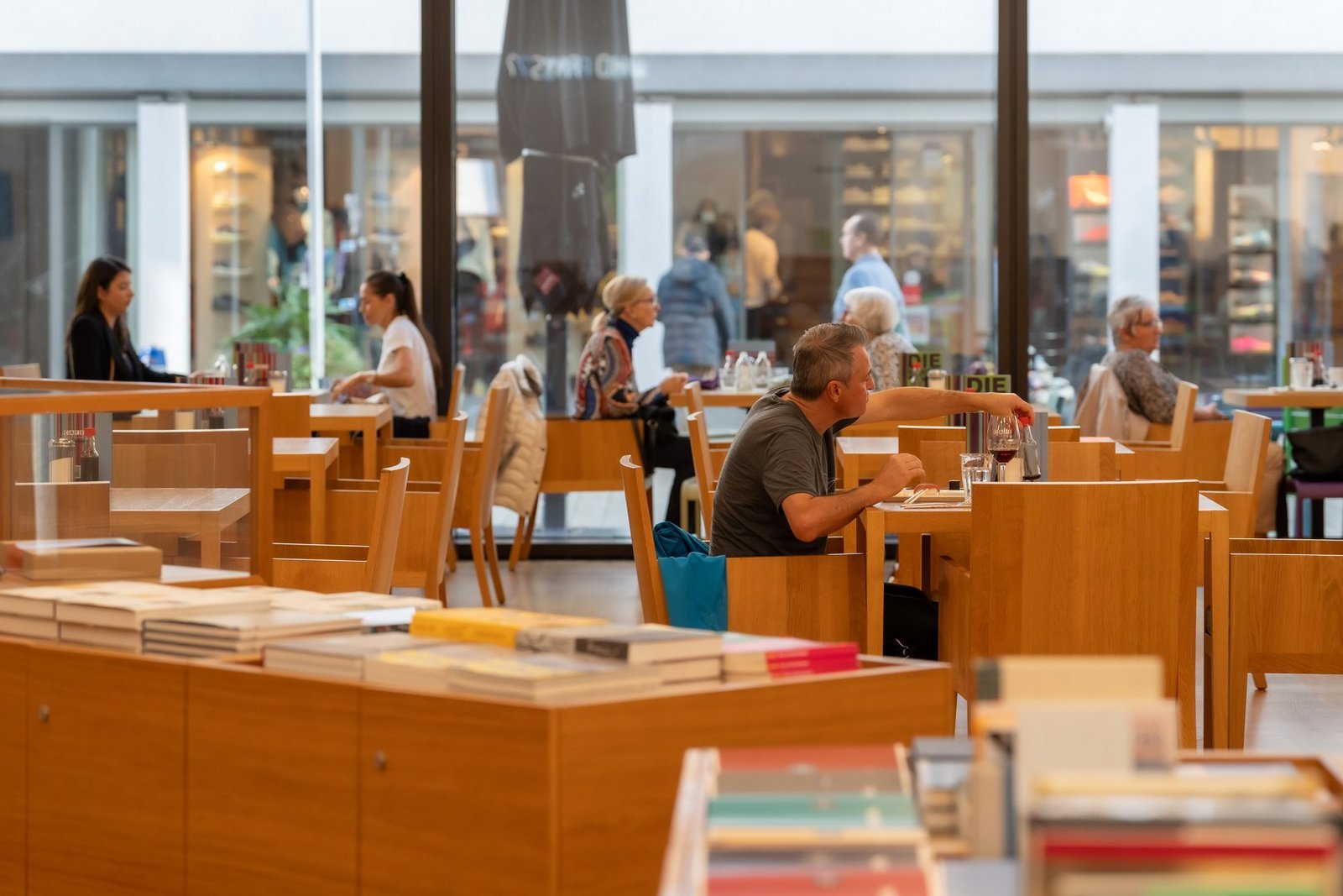 Gut besuchtes Museums-Café mit hellen Holzmöbeln und großen Glasfronten, im Vordergrund sind Büchertische mit Katalogen aufgestellt.