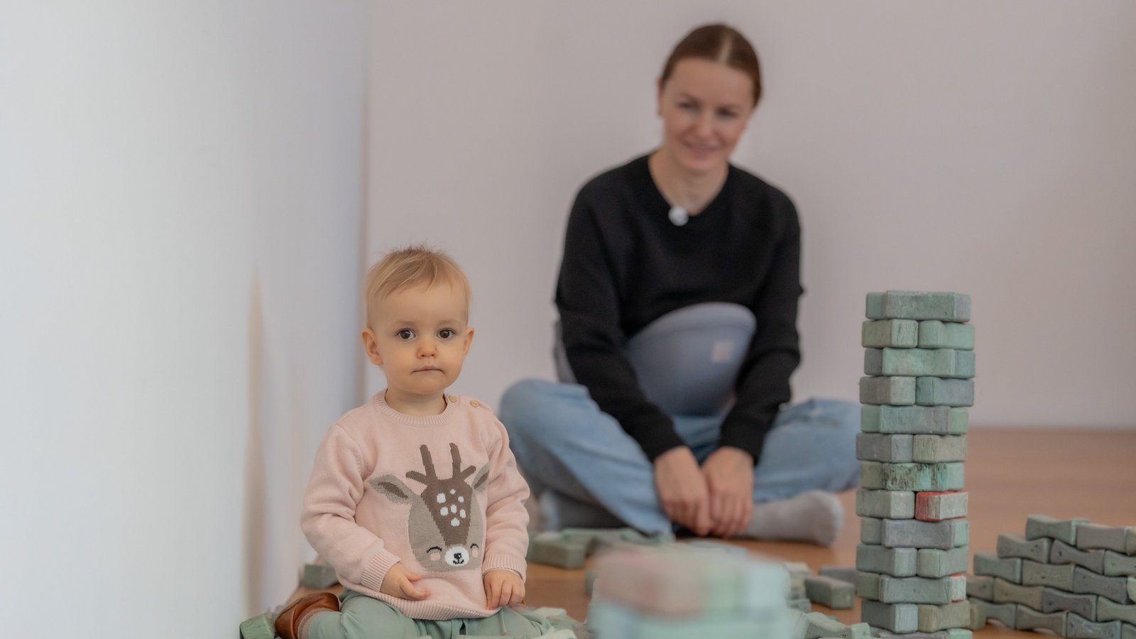 Kleinkind beim Spielen mit Bausteinen auf dem Boden, im Hintergrund eine Frau.