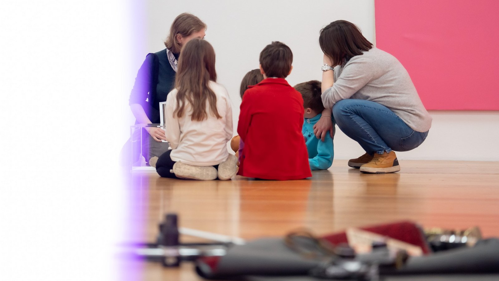 Eine Gruppe von Kindern sitzt zusammen mit zwei erwachsenen Frauen im Kreis auf dem Holzboden des Kunstmuseums.